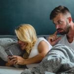 Young man is watching his girlfriends phone over her shoulder while they are laying in bed (istockphoto)