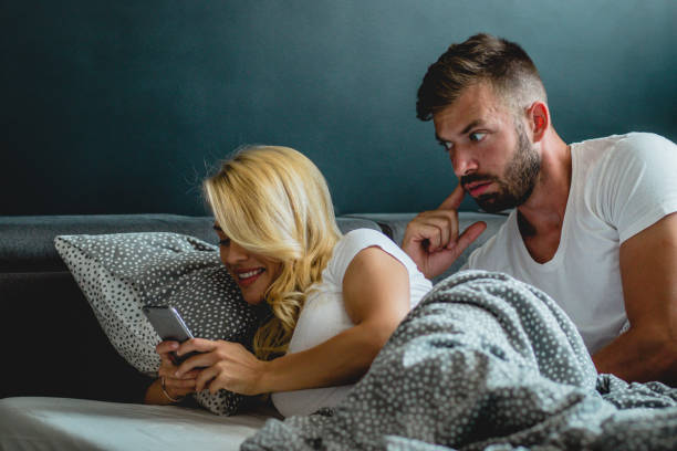 Young man is watching his girlfriends phone over her shoulder while they are laying in bed (istockphoto)